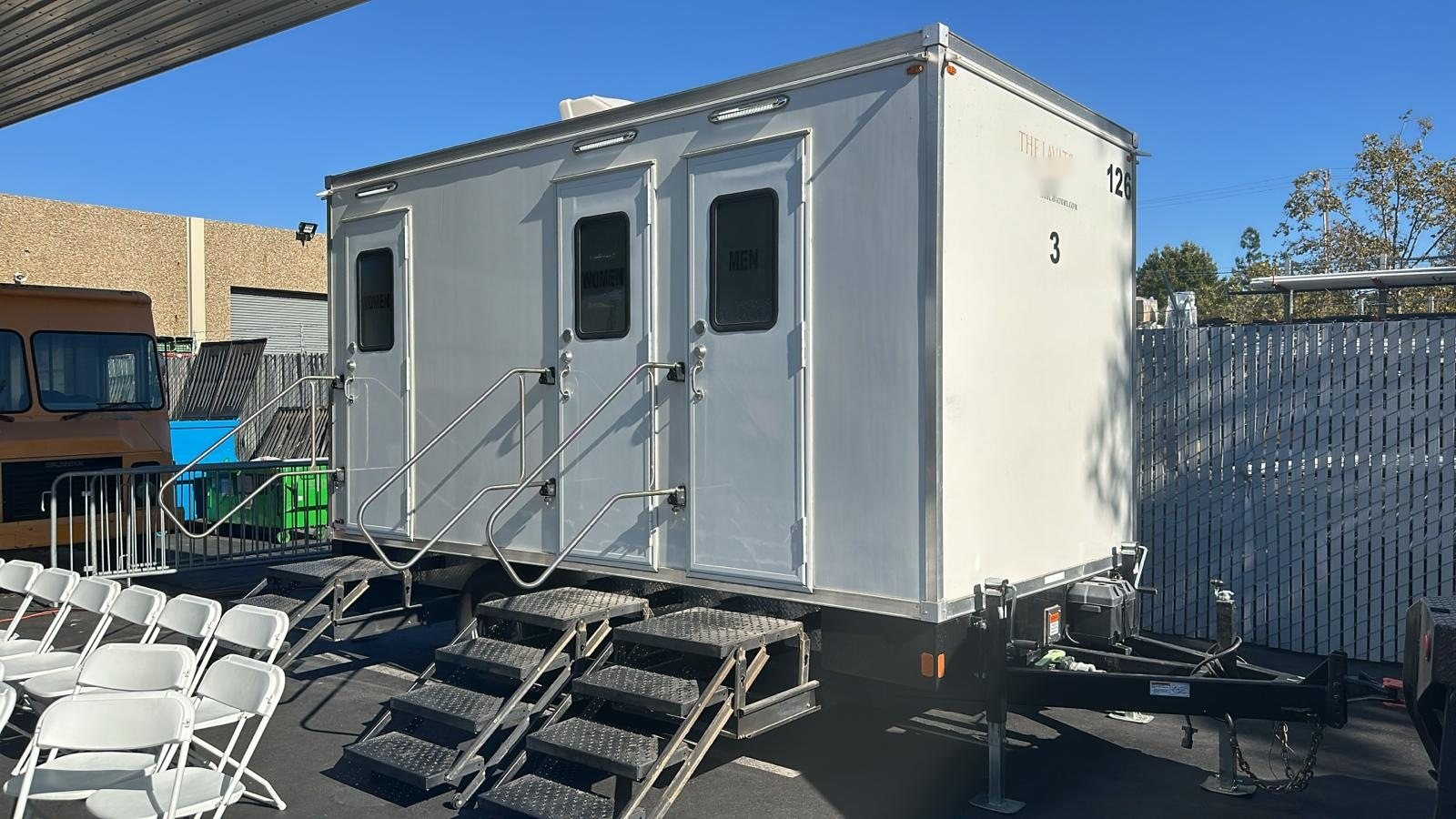 A Lavatory 3-Station restroom trailer with two women's doors and one men's door next to rows of white folding chairs at an event