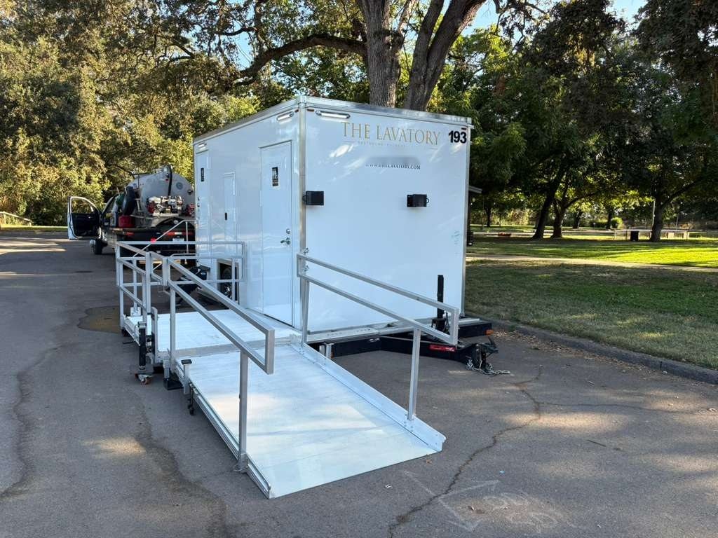 A Lavatory ADA restroom trailer with an aluminum ramp and rails parked beside a shaded park lawn