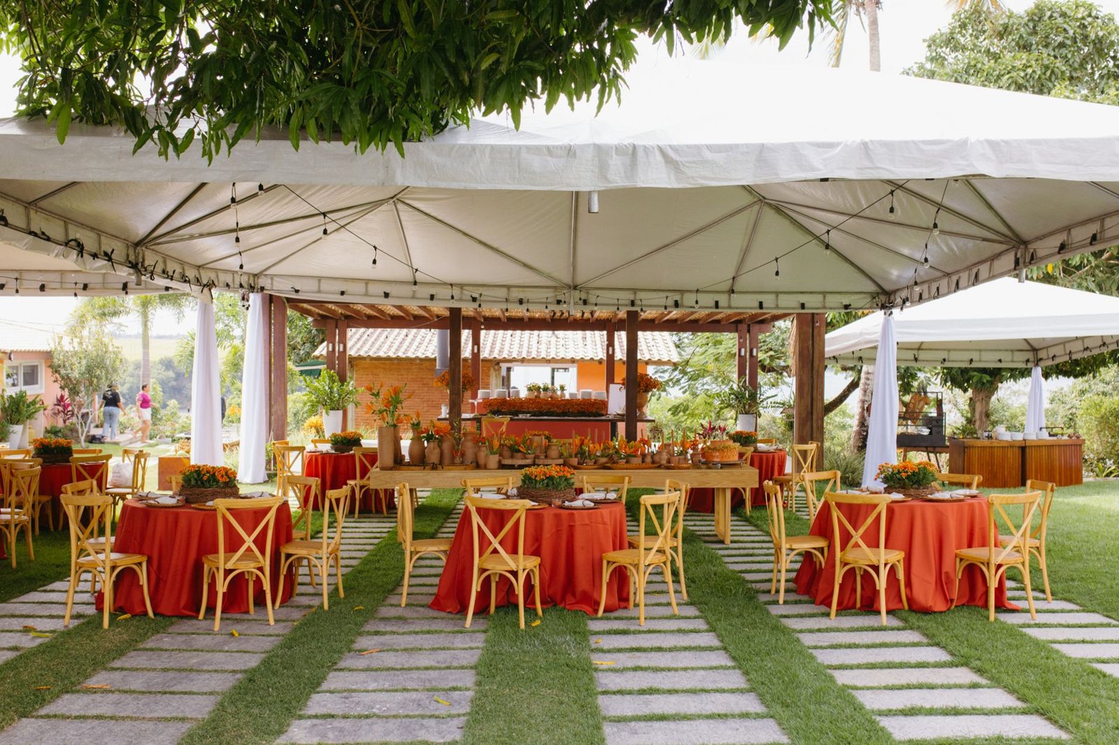 An outdoor reception under white tents with red tablecloths and wooden chairs on a lawn