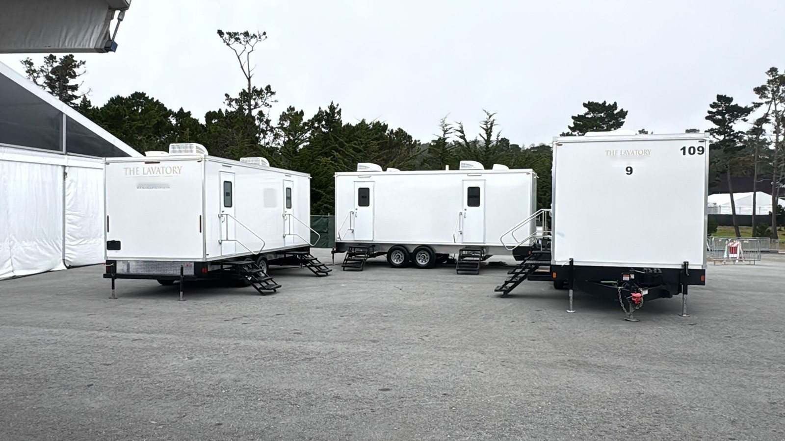 Three Lavatory restroom trailers, including a 9-Station, set on a paved event lot beside a white hospitality tent