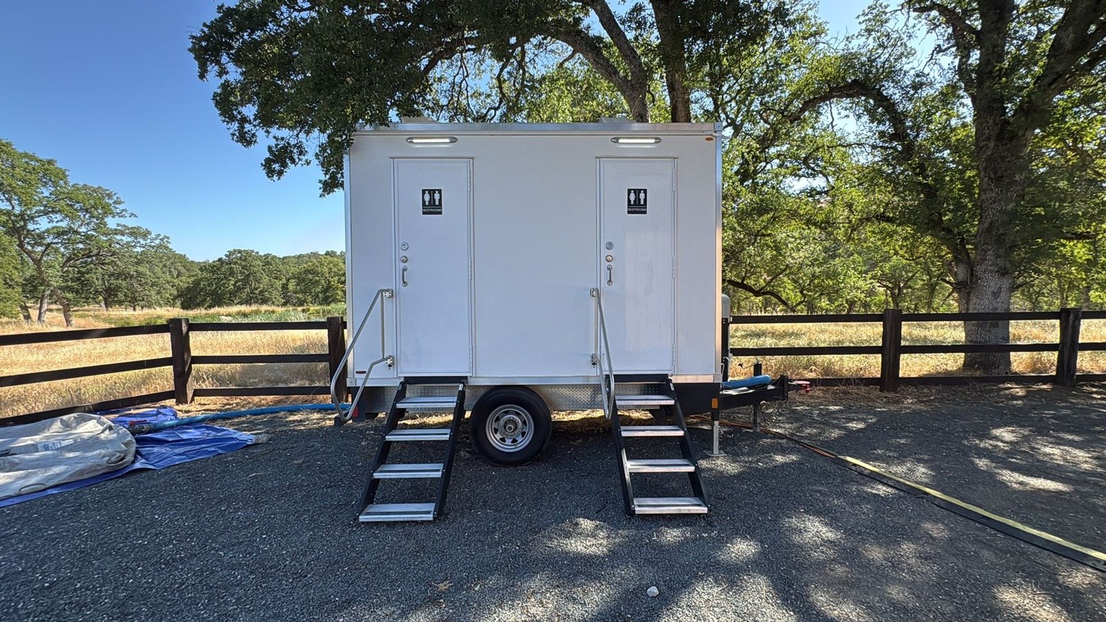 A Lavatory 2-Station restroom trailer with steel stairs on a gravel pad beside a wooden ranch fence under big oak trees