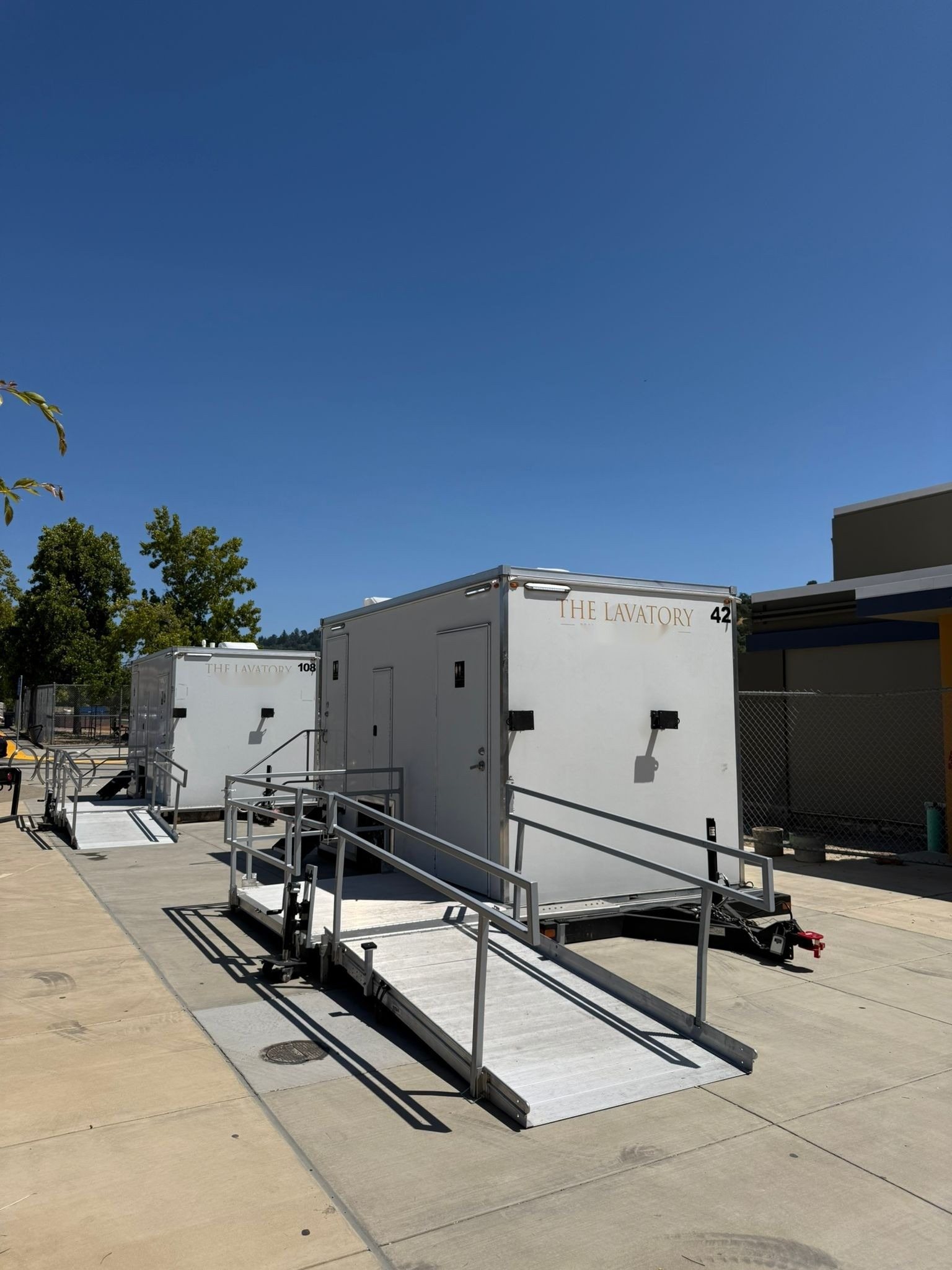 Two Lavatory ADA restroom trailers with aluminum ramps and handrails on a school campus walkway under a clear sky