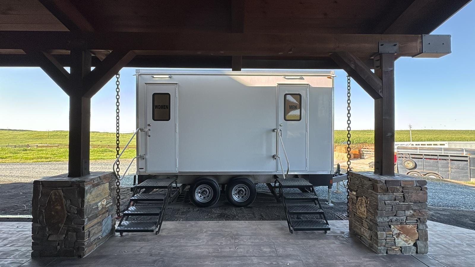 A Lavatory 2-Station luxury restroom trailer with separate women's and men's doors and steel stairs, parked under a timber pavilion with stone pillars at a ranch event site