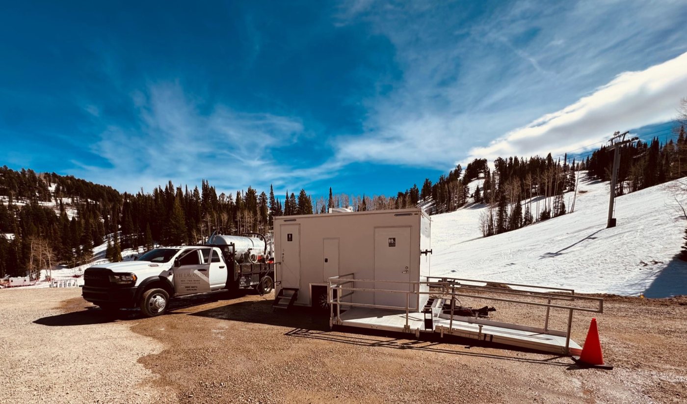 A Lavatory restroom trailer with an accessible ramp set on a gravel lot at a ski area, with snow on the slopes and a service truck beside it