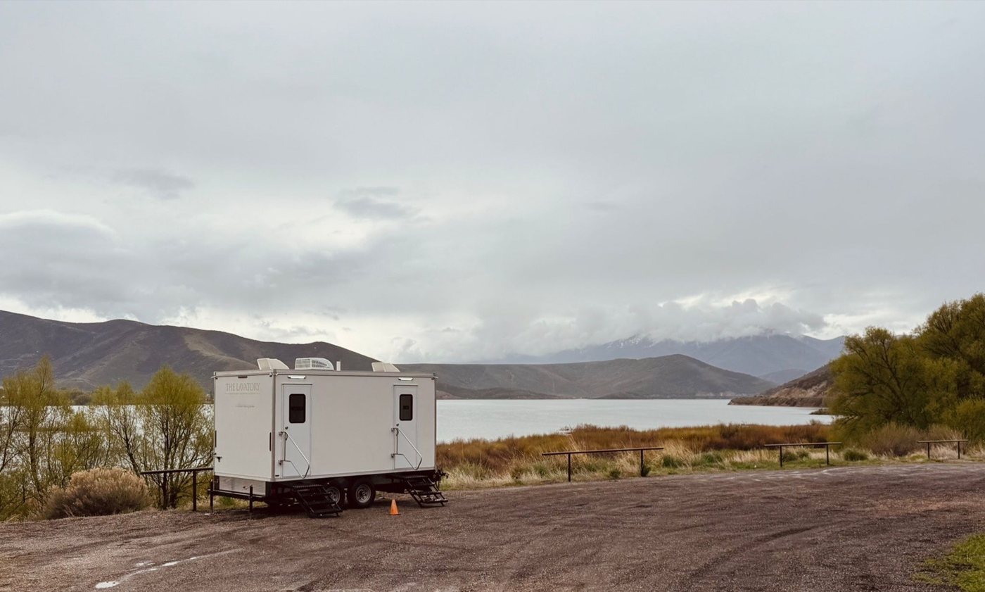 A Lavatory restroom trailer parked on a gravel lot beside a mountain reservoir under a cloudy sky