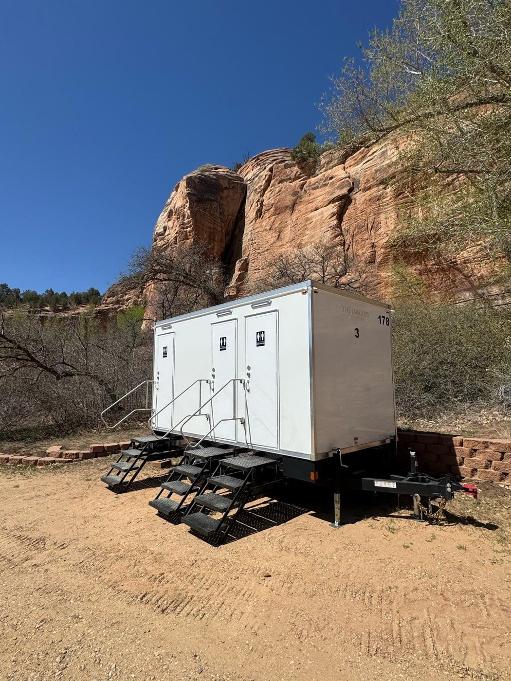 A Lavatory 3-Station restroom trailer on a dirt pad at the foot of a red sandstone cliff, with stairs deployed at each door