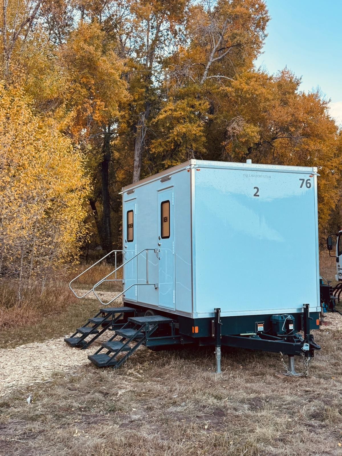 A Lavatory 2-Station bathroom trailer set on gravel among trees with yellow fall leaves