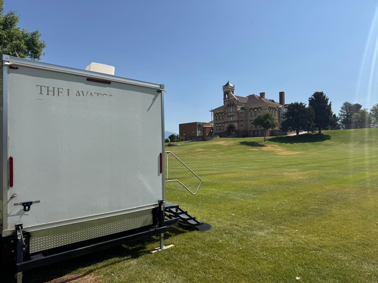A Lavatory restroom trailer on a wide lawn with a historic brick building in the distance