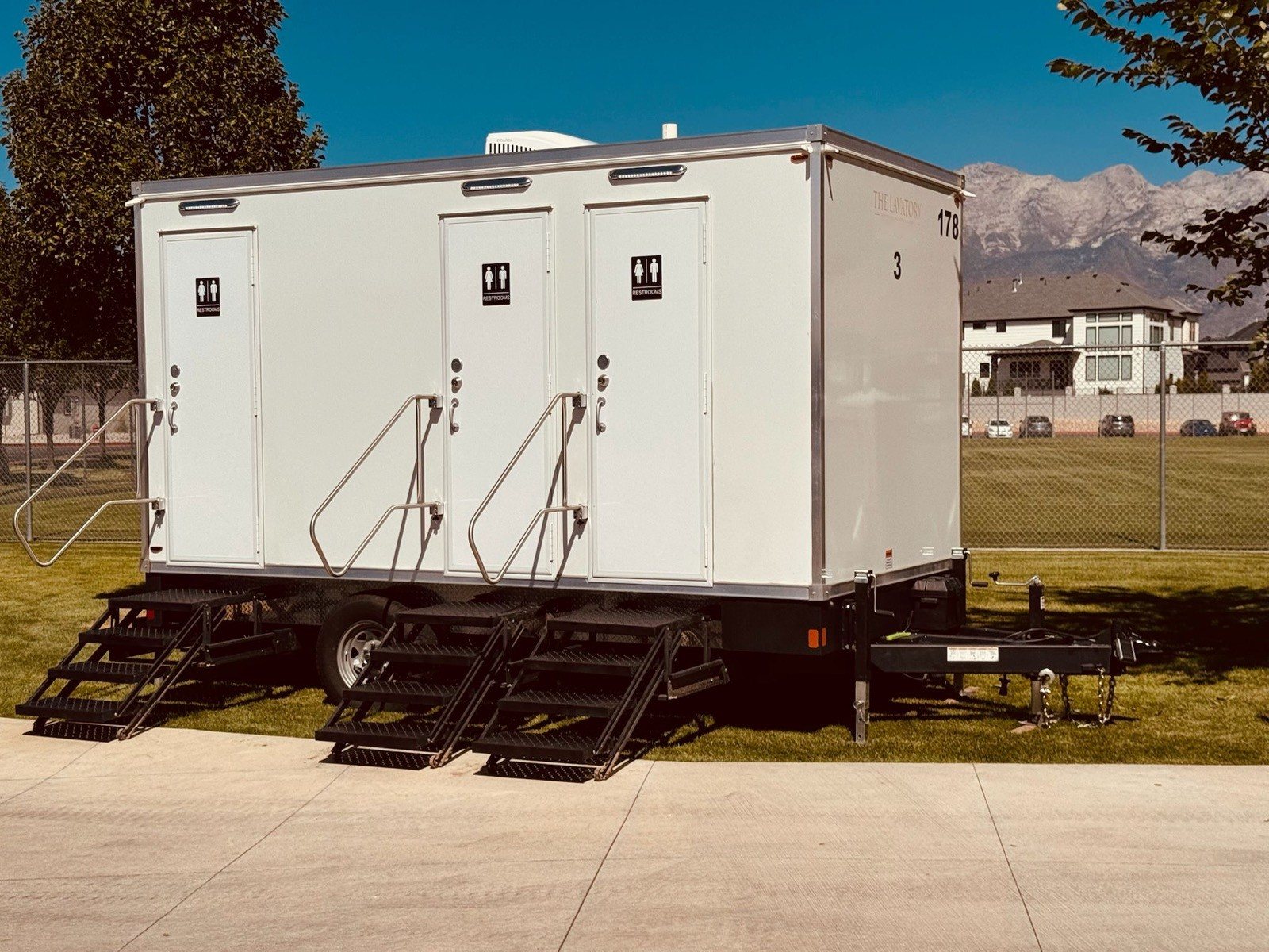 A Lavatory 3-Station restroom trailer parked on a sidewalk edge beside a fenced lawn, with homes and mountains behind it