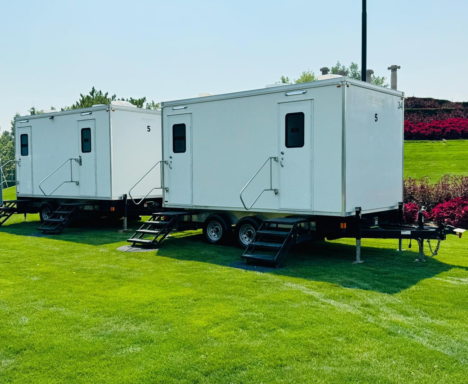 Two Lavatory 5-Station restroom trailers parked side by side on a green lawn with red flowering shrubs behind them