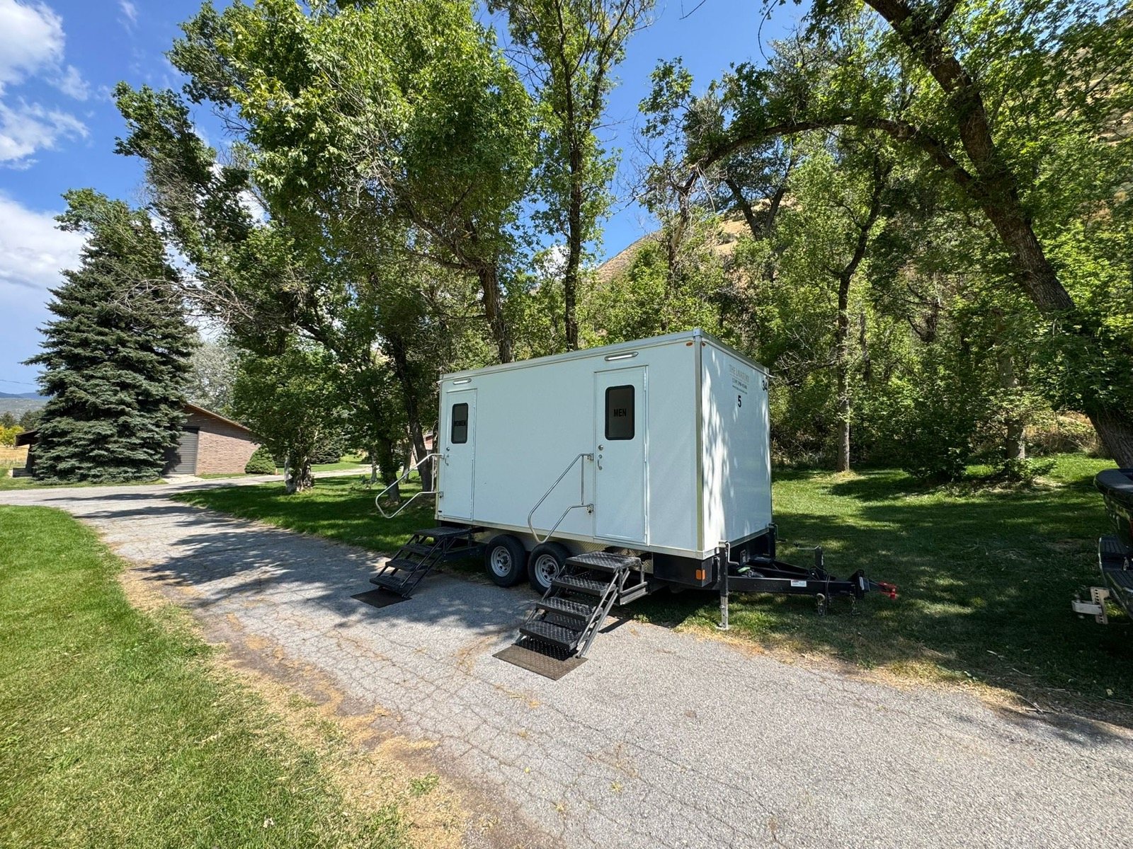 A Lavatory restroom trailer on a tree-lined gravel drive beside a lawn, with its steps and handrail deployed