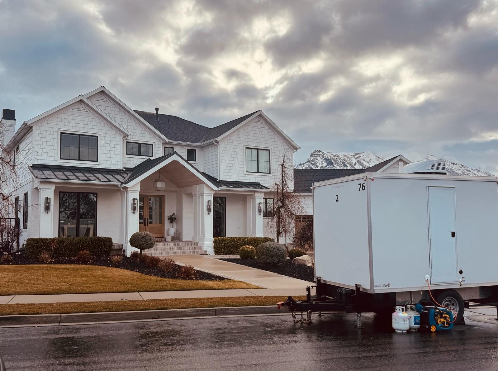 A Lavatory 2-Station restroom trailer at the curb in front of a large white house, with a generator and propane tanks beside it