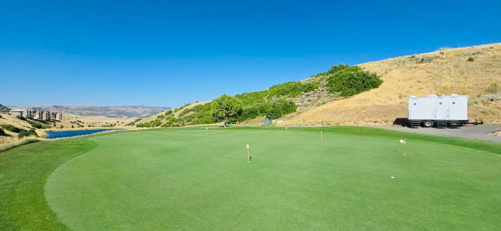A Lavatory 3-Station restroom trailer parked on a paved path beside a golf putting green below a dry hillside