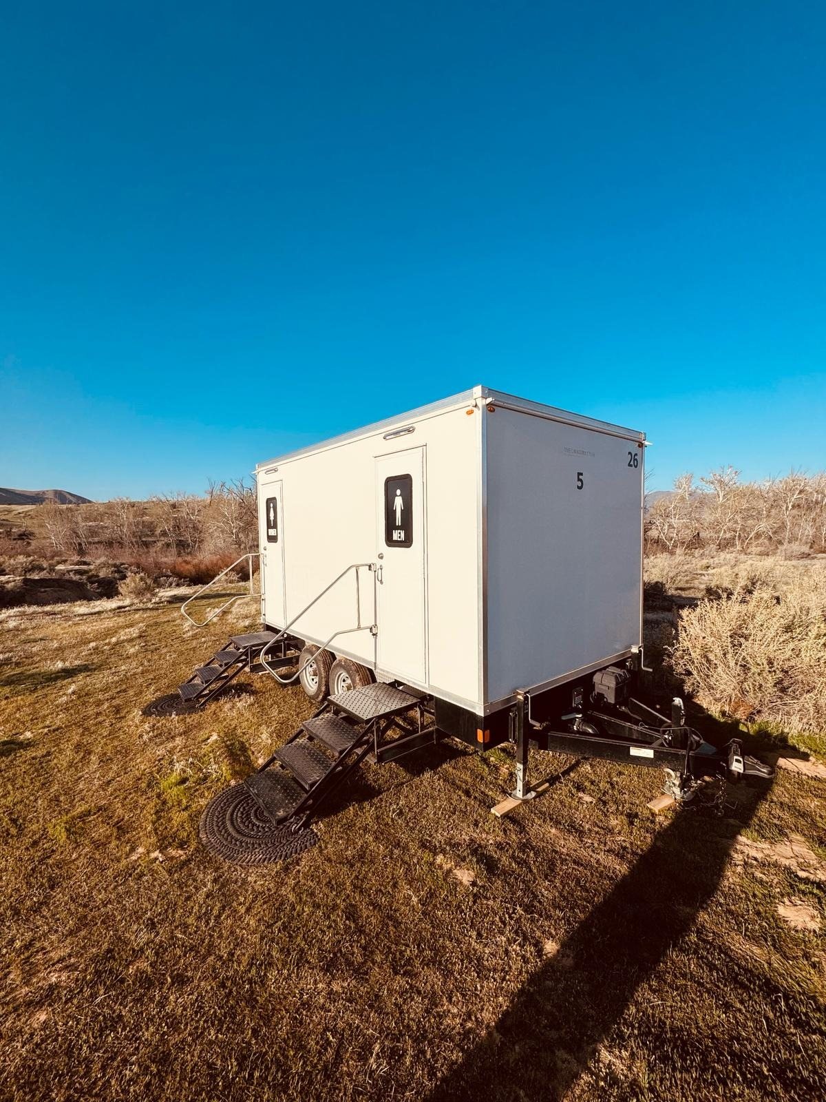 A Lavatory 5-Station restroom trailer set on blocks on open sagebrush ground, with stairs at the men's and women's doors