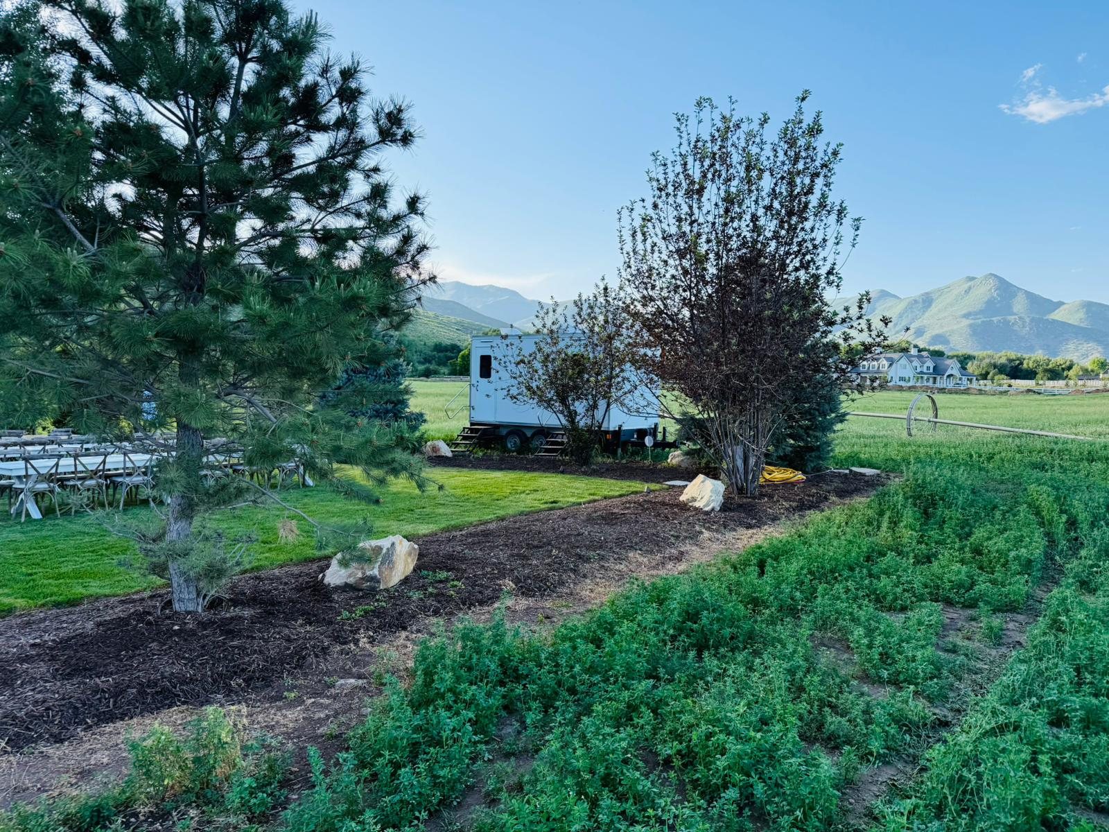 A Lavatory restroom trailer tucked behind trees next to a lawn set with long farm tables and wood chairs, mountains behind