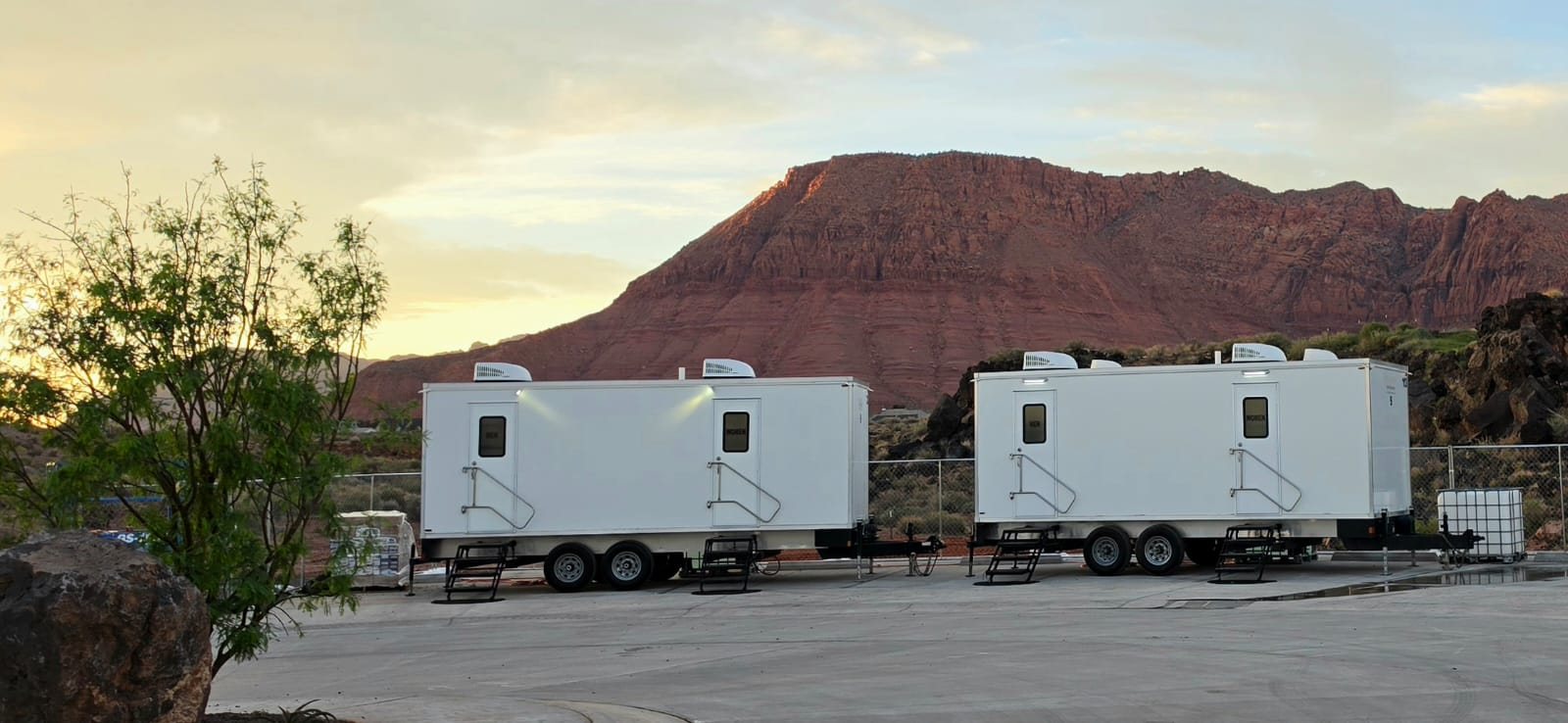 Two Lavatory restroom trailers parked on a paved lot below red rock cliffs at sunrise