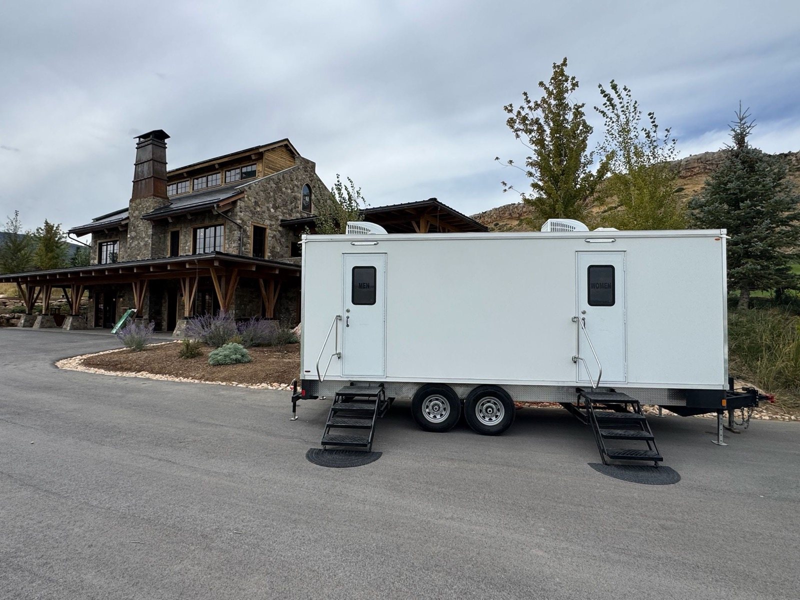 A Lavatory 9-Station restroom trailer on a paved drive in front of a stone and timber lodge