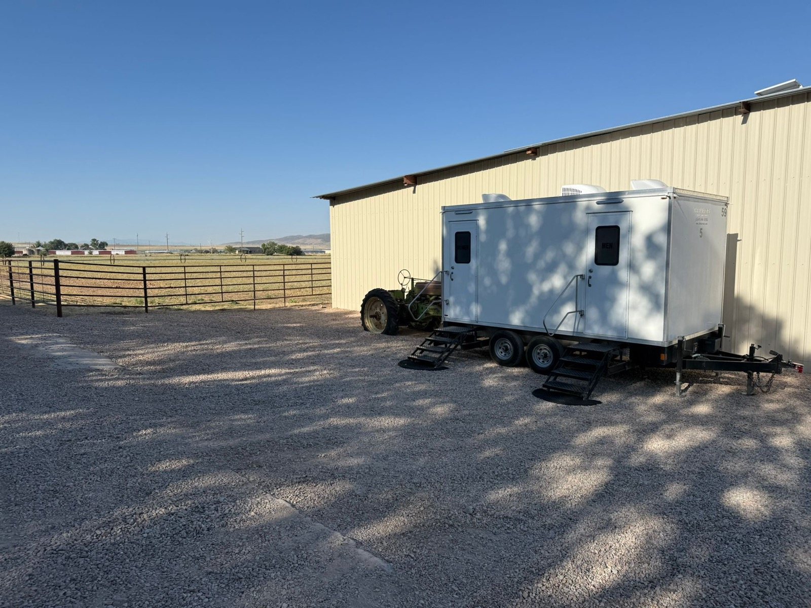 A Lavatory restroom trailer parked on gravel beside a metal barn and fenced pasture