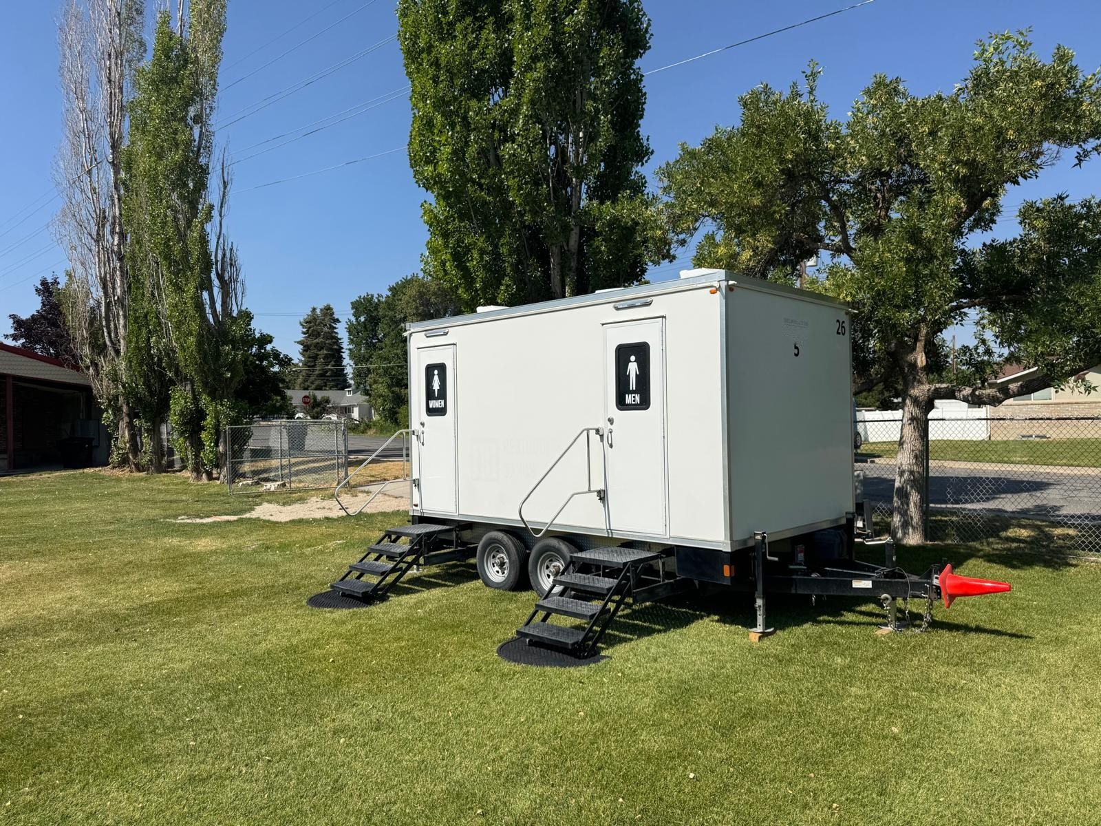 A Lavatory 2-Station restroom trailer on a green lawn under tall trees, with stairs at each door