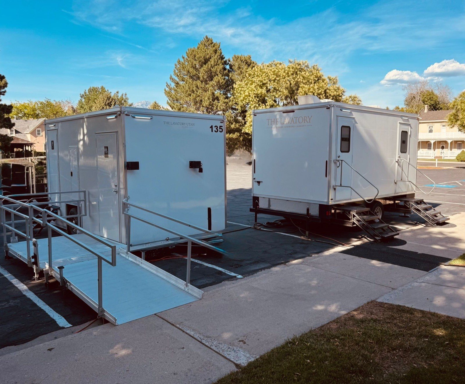 Two Lavatory restroom trailers in a parking lot, one with a metal wheelchair ramp and handrails