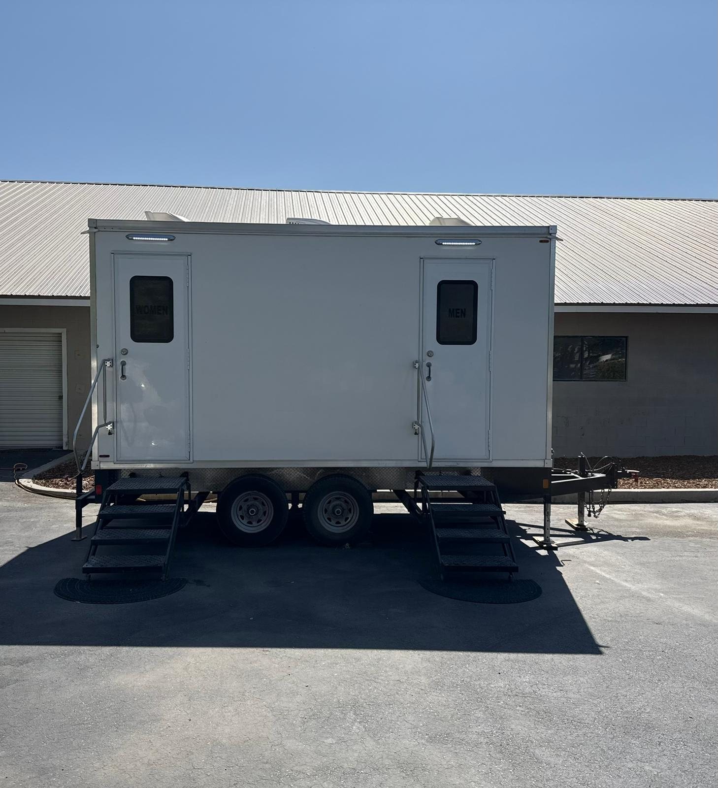 A Lavatory 2-Station bathroom trailer on asphalt in front of a metal-sided building