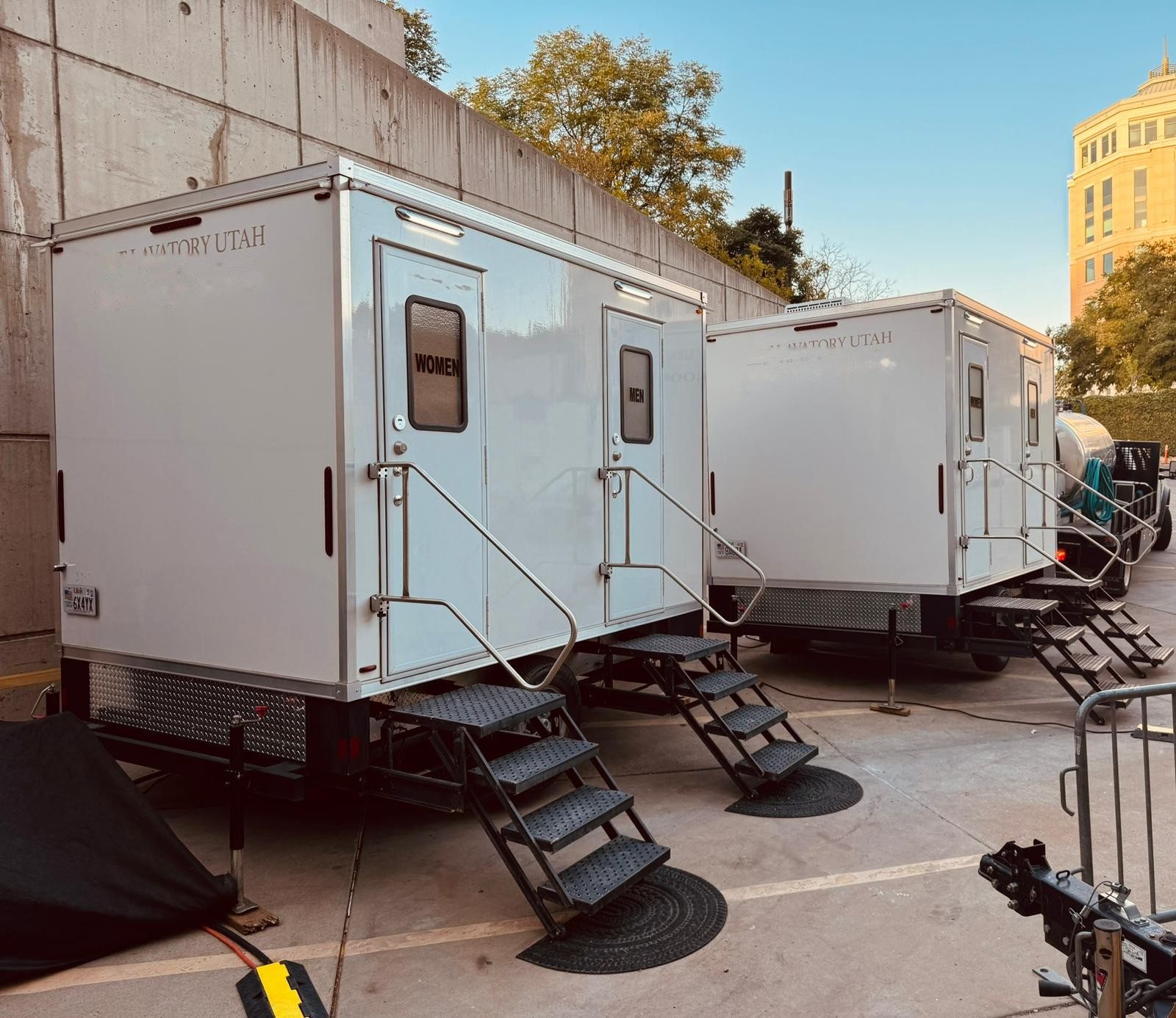 Two Lavatory restroom trailers parked side by side against a concrete wall outside an office building