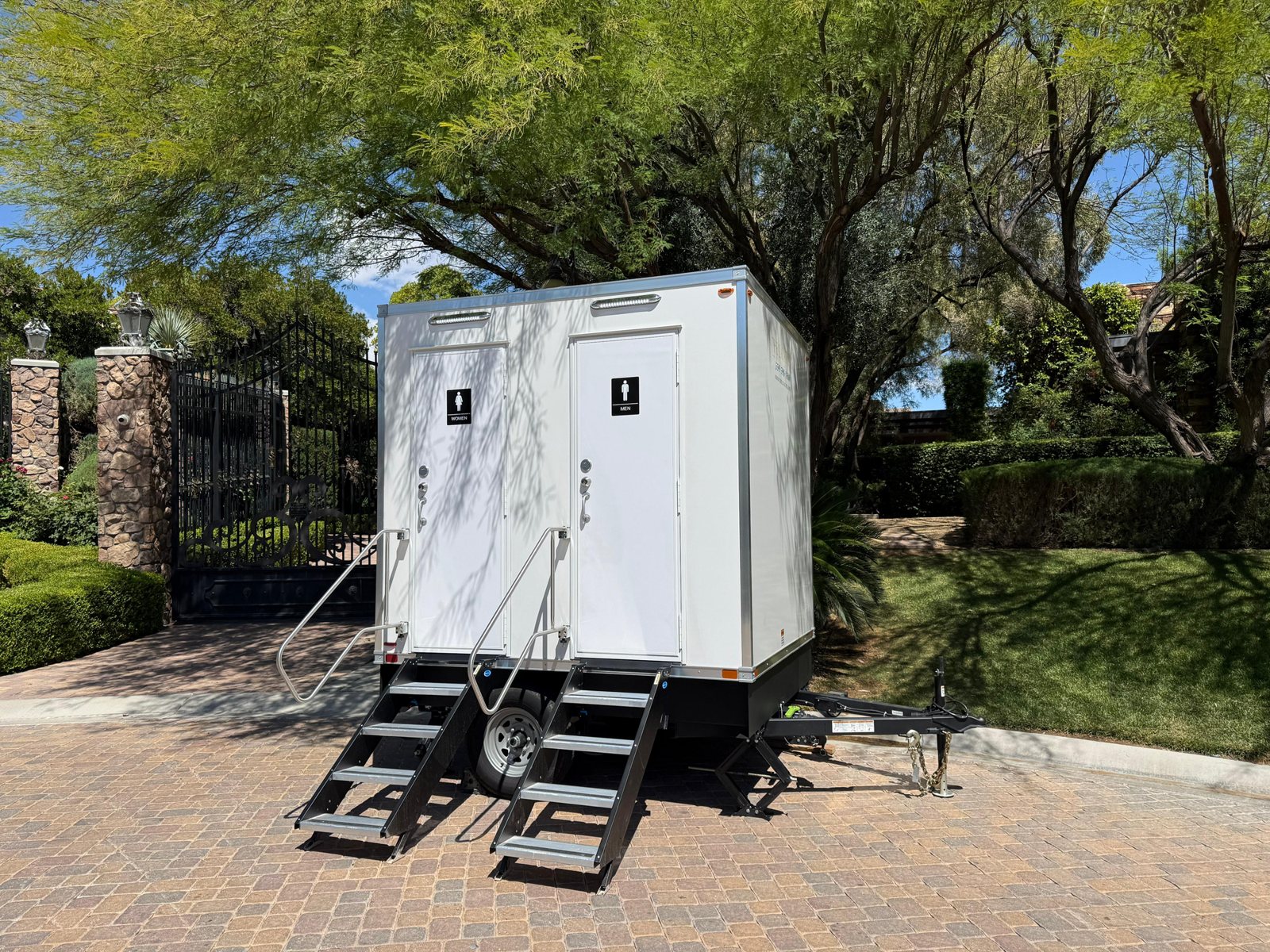 A two station Lavatory restroom trailer set on a paver driveway in front of a gated estate, under desert shade trees