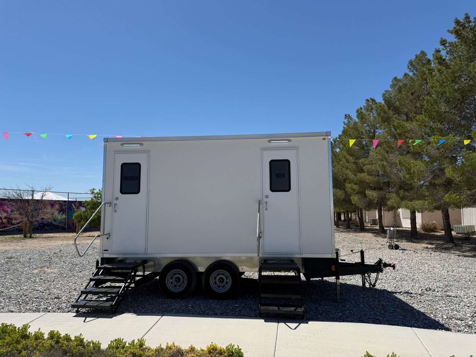 A Lavatory restroom trailer with women's and men's doors set on gravel under strings of festival flags