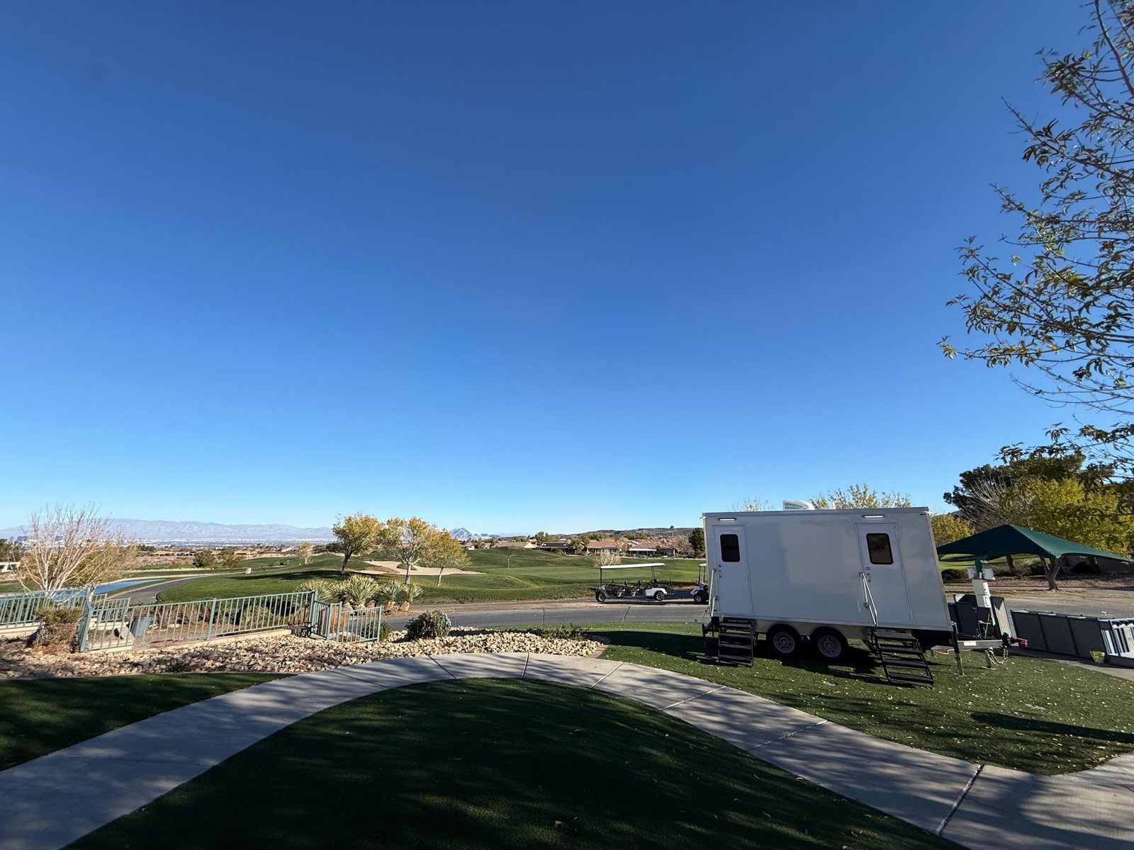 A Lavatory restroom trailer parked on turf at the edge of a desert golf course, with carts and the valley behind it