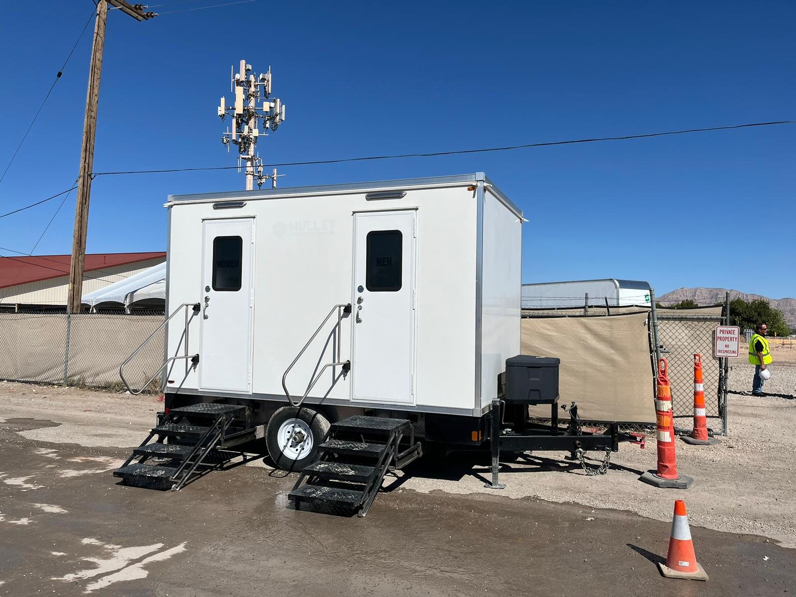 A worker in a hi-vis vest stands by the fence near a Lavatory restroom trailer set on a gravel work lot with traffic cones