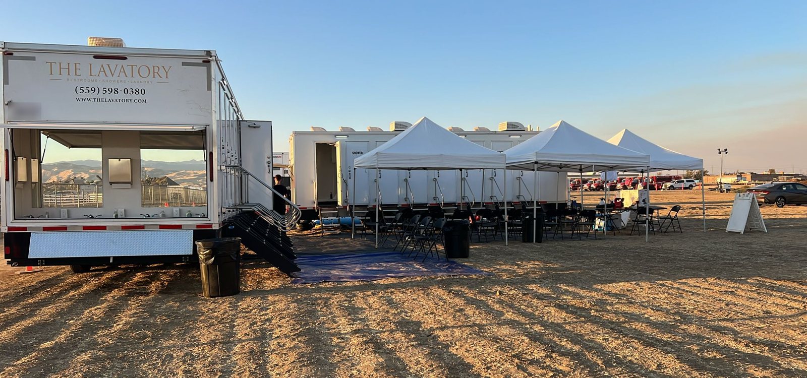 Shower trailer set up behind three white canopy tents with folding chairs on a dry dirt lot at sunset, a Lavatory trailer with its sink side open in front