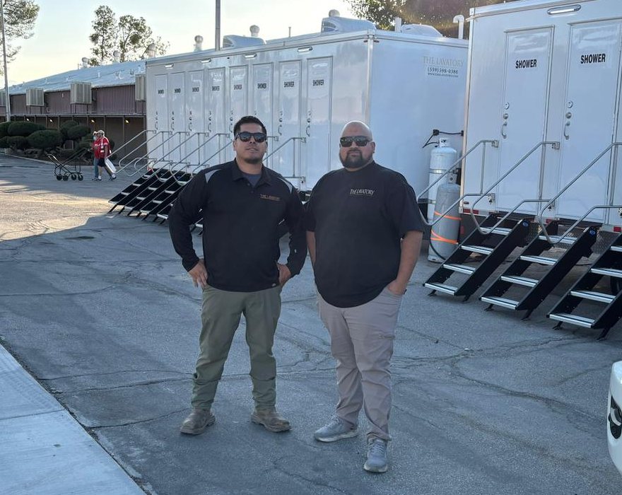 Two Lavatory crew members in black shirts standing on a paved lot beside two shower trailers with stairs at the doors