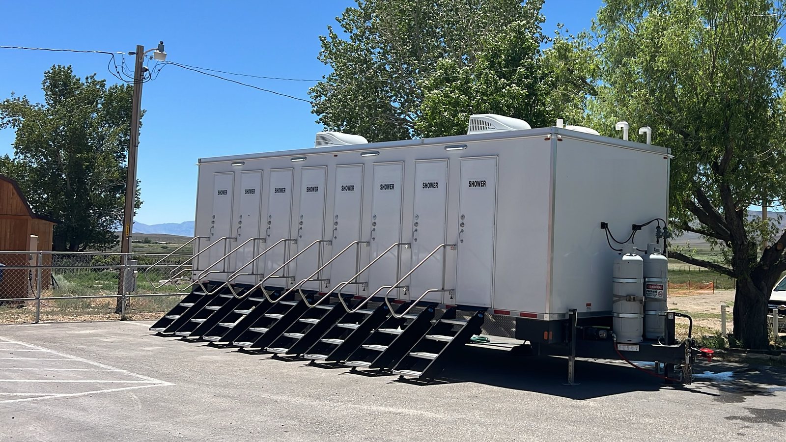 8-station shower trailer with a stair at each of its eight doors, parked on a paved lot with propane tanks on the tongue and dry desert mountains behind