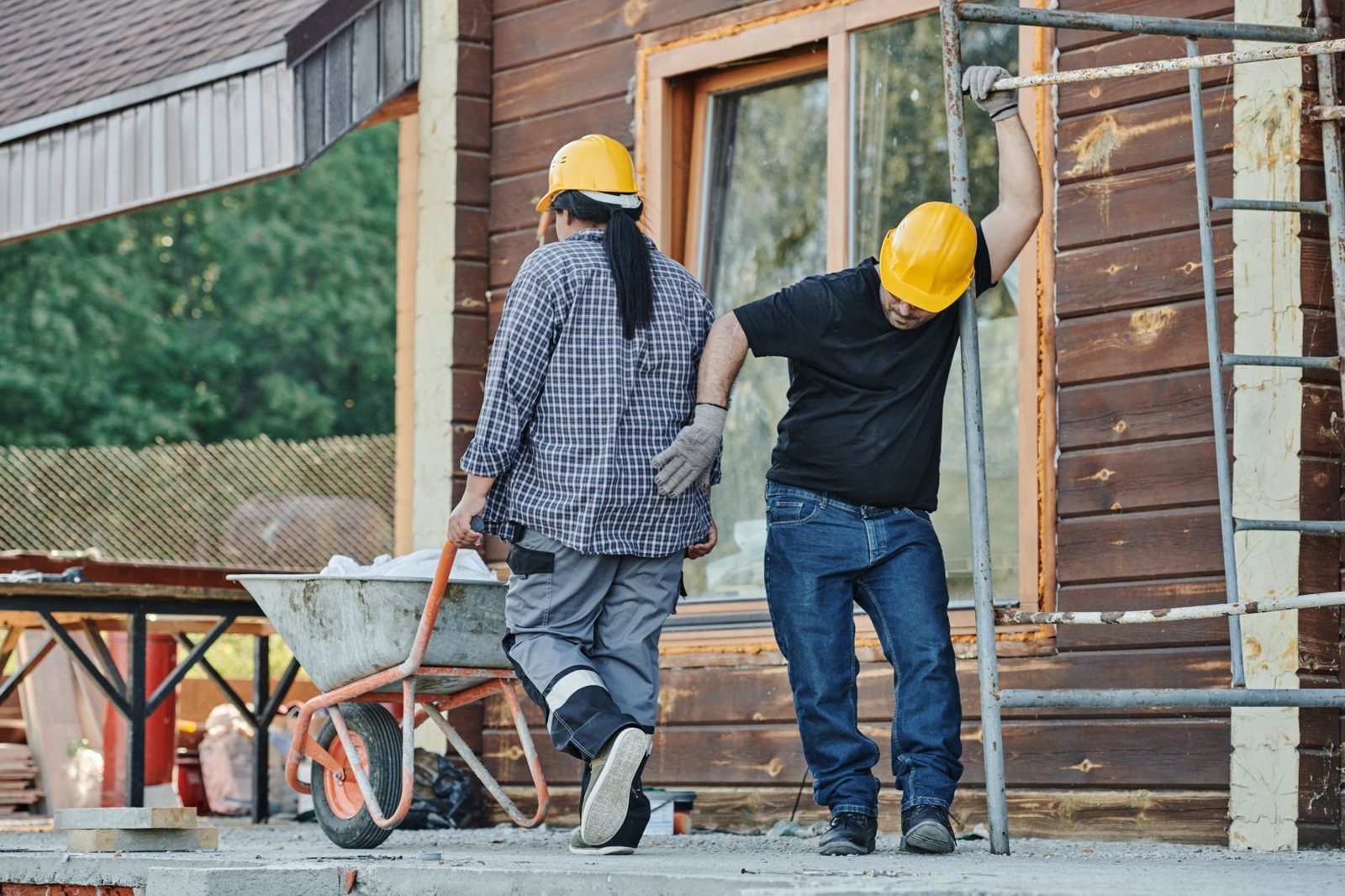 Two construction workers in yellow hard hats move materials beside scaffolding on a building renovation