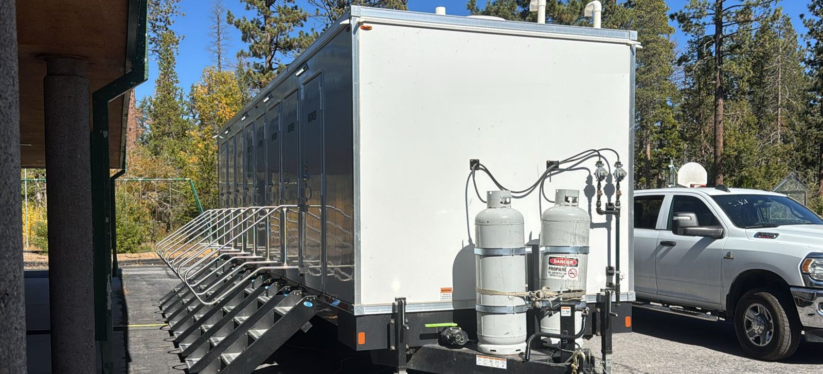 8-Station private shower trailer with stairs at each door and twin propane tanks on the tongue, parked on an asphalt lot under tall pines