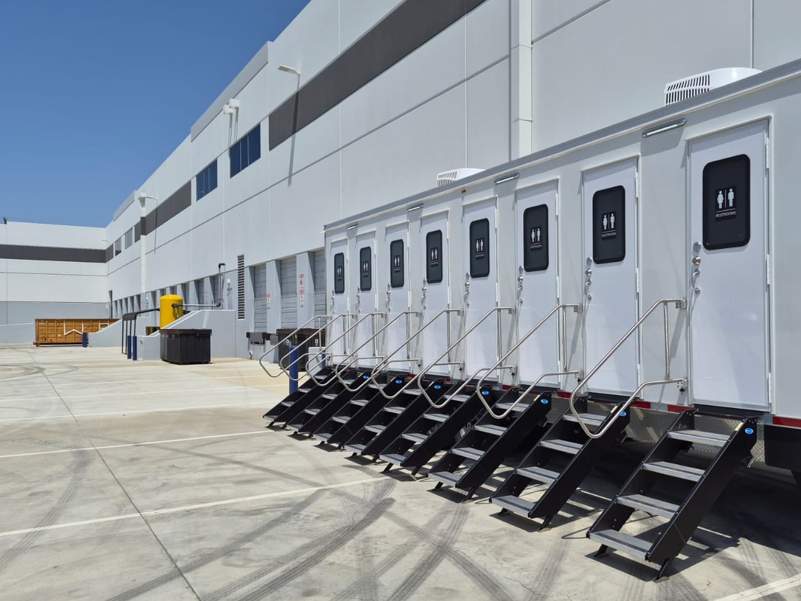 A long Lavatory private stall restroom trailer with a row of doors and steps set along the loading dock of a large warehouse