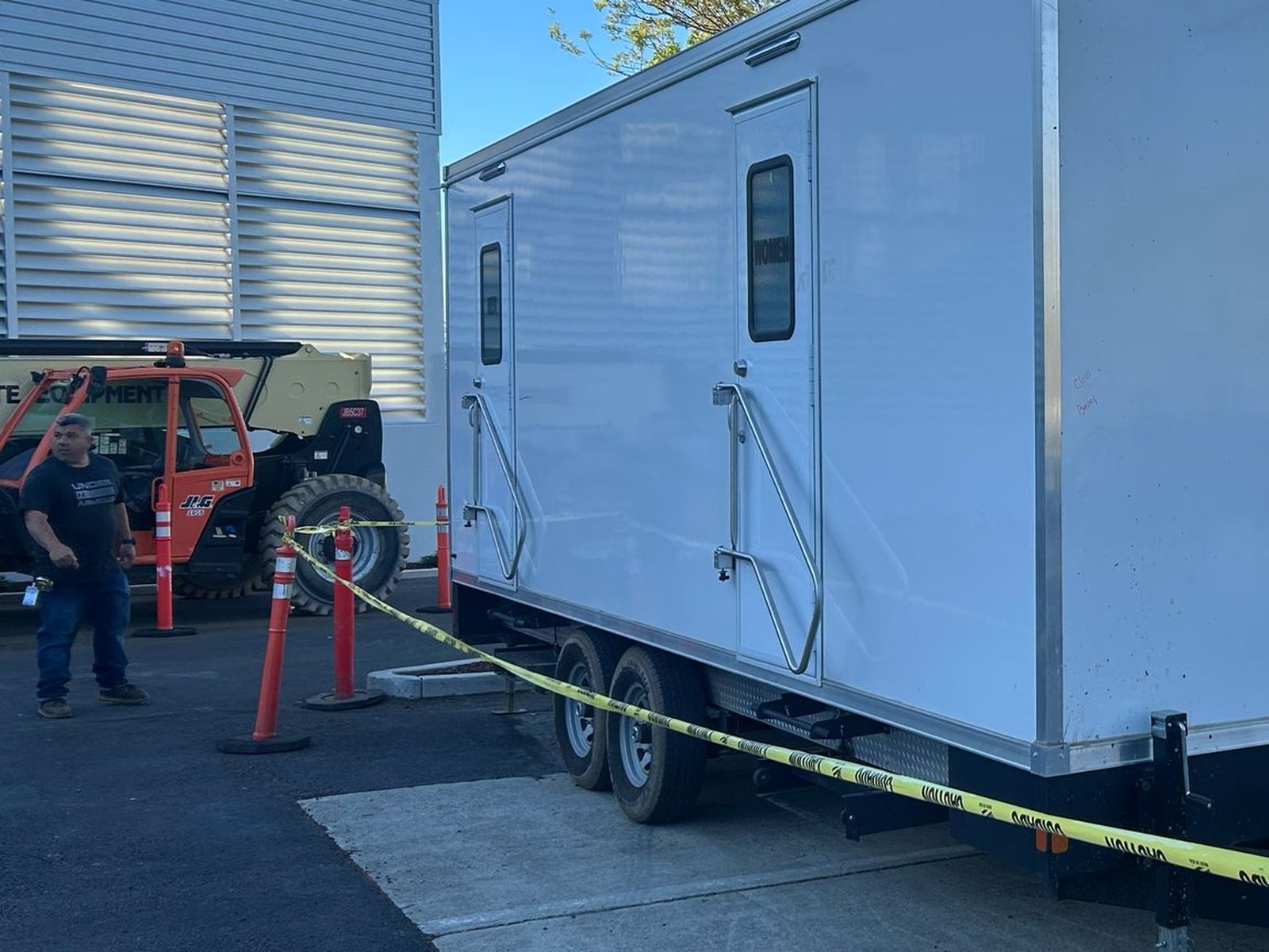 A worker stands by a telehandler next to a Lavatory restroom trailer set behind caution tape in a construction yard
