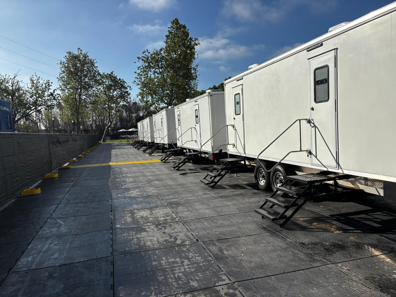 A row of Lavatory restroom trailers lined up on ground mats along a fenced event lane