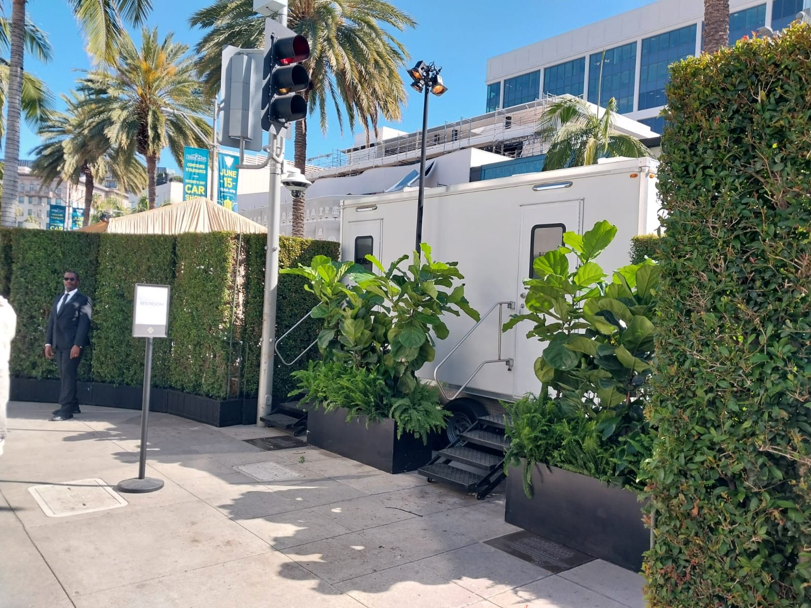 An event staff member in a dark suit stands at a hedge wall entrance beside a Lavatory restroom trailer dressed with potted plants