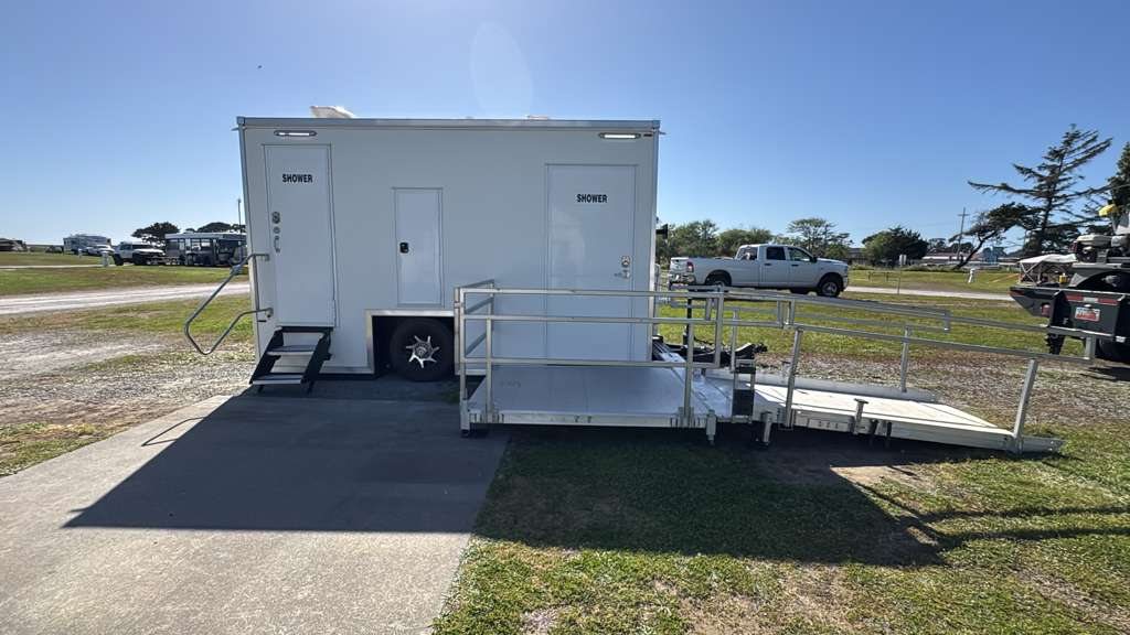 ADA + 2 private shower trailer with its accessible ramp and railings deployed, parked on a paved pad at the edge of a grass field