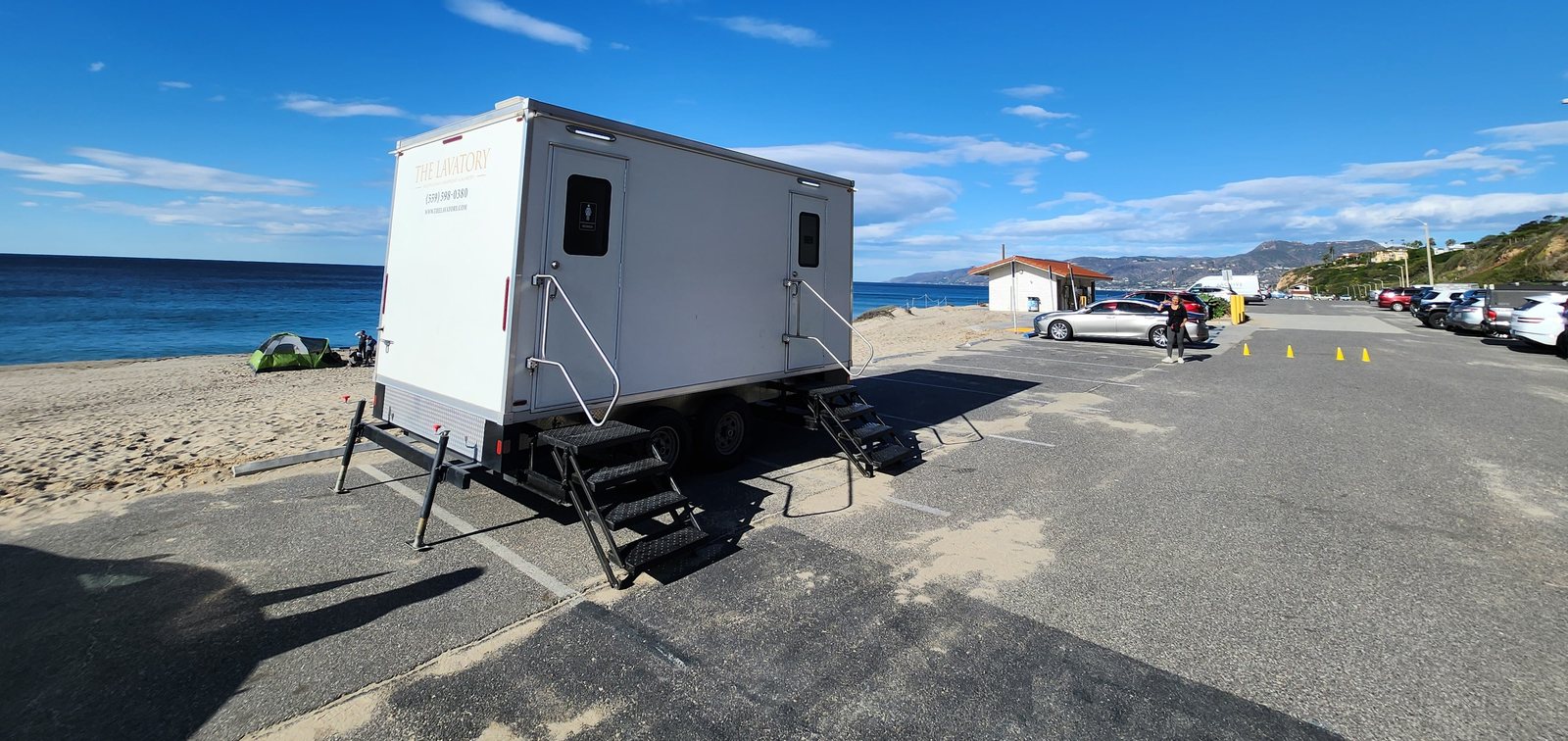 2-station shower and restroom combo trailer set on a paved beach parking lot, with the ocean and coastal hills behind it