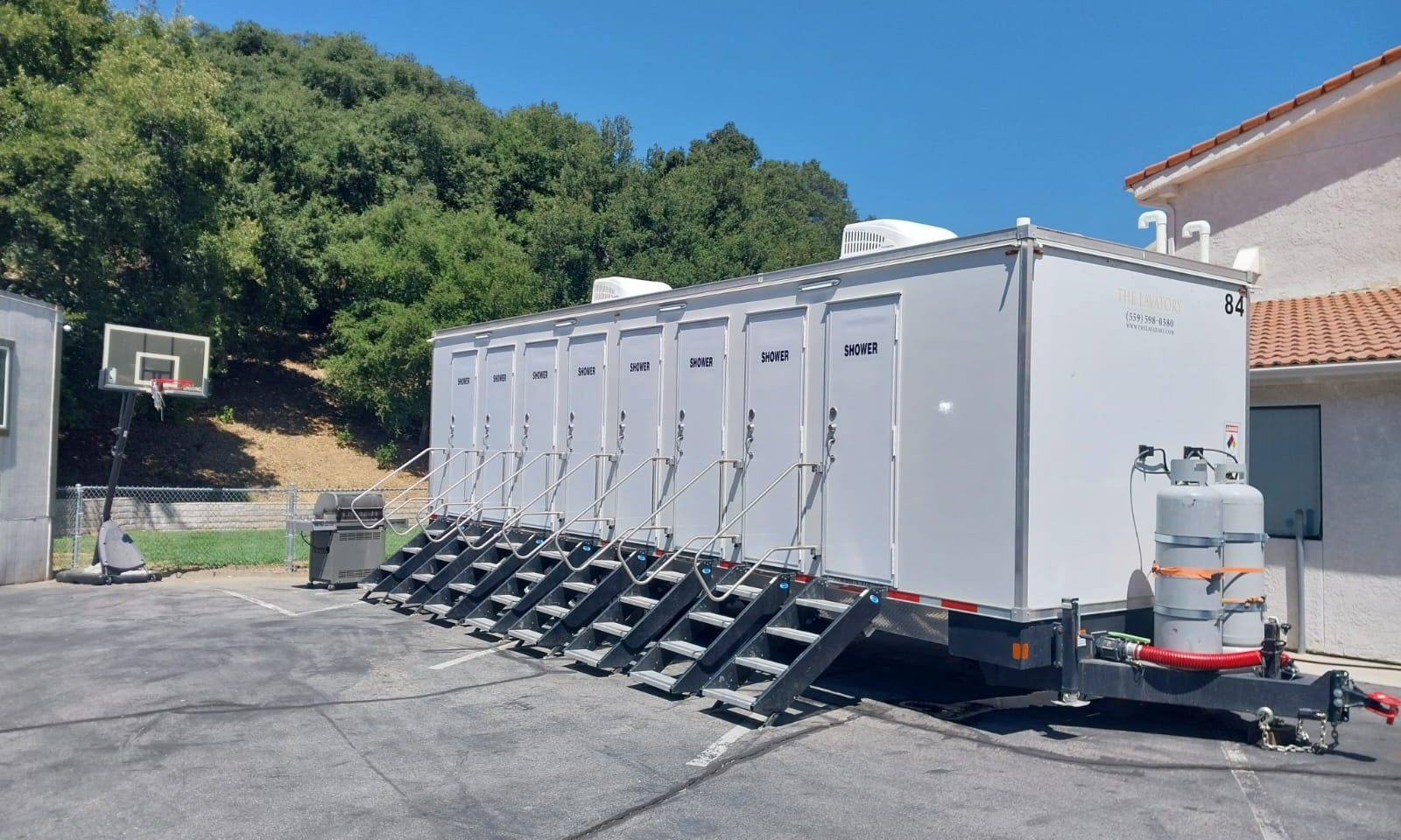 8-station private shower trailer with a stair at each of its eight doors, parked on a paved campus lot beside a basketball hoop, a tile-roofed building and oak-covered hills