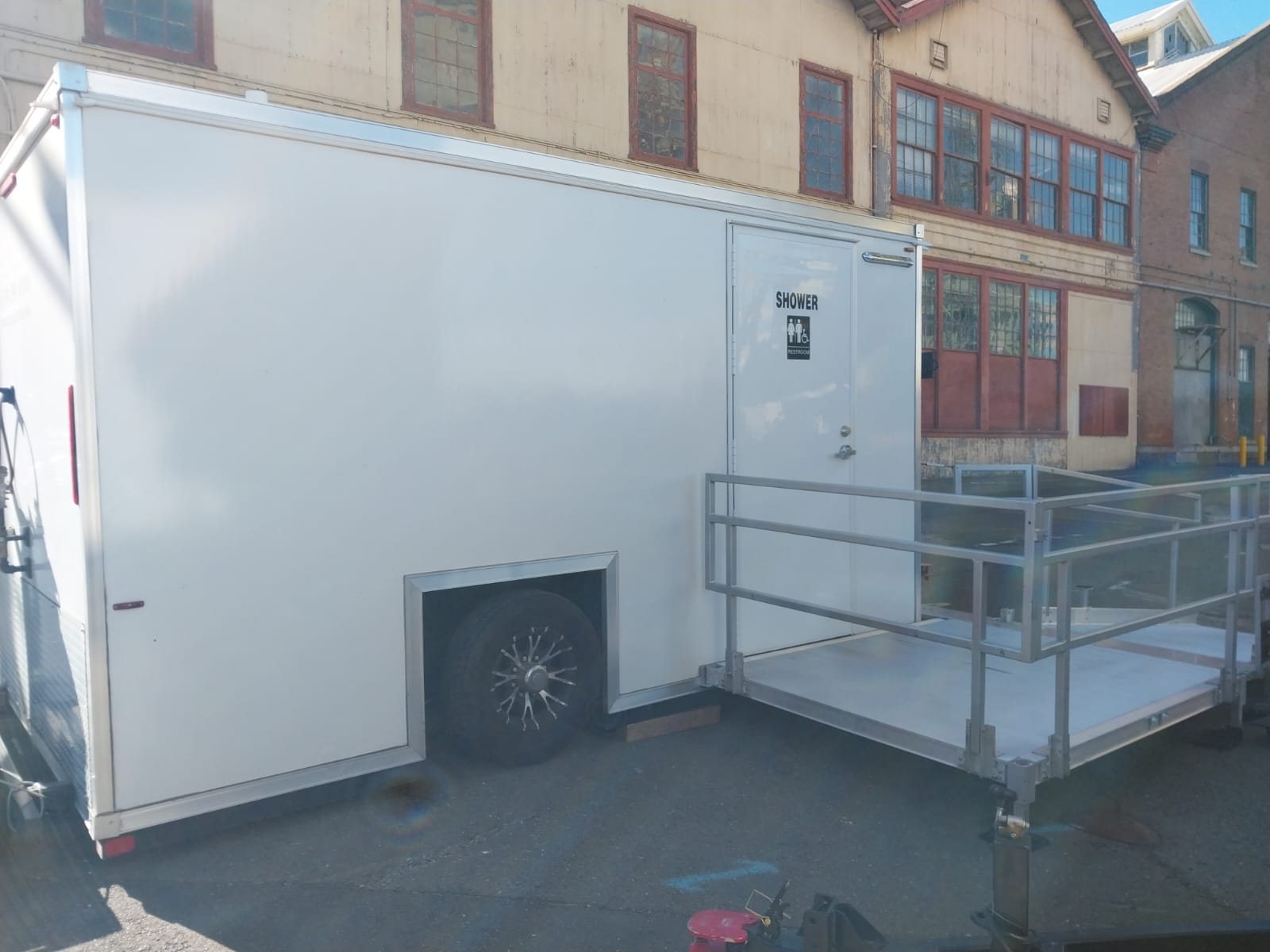 ADA plus 2 shower trailer with a SHOWER door and an aluminum ramp platform, parked on an asphalt yard beside an old brick and stucco industrial building
