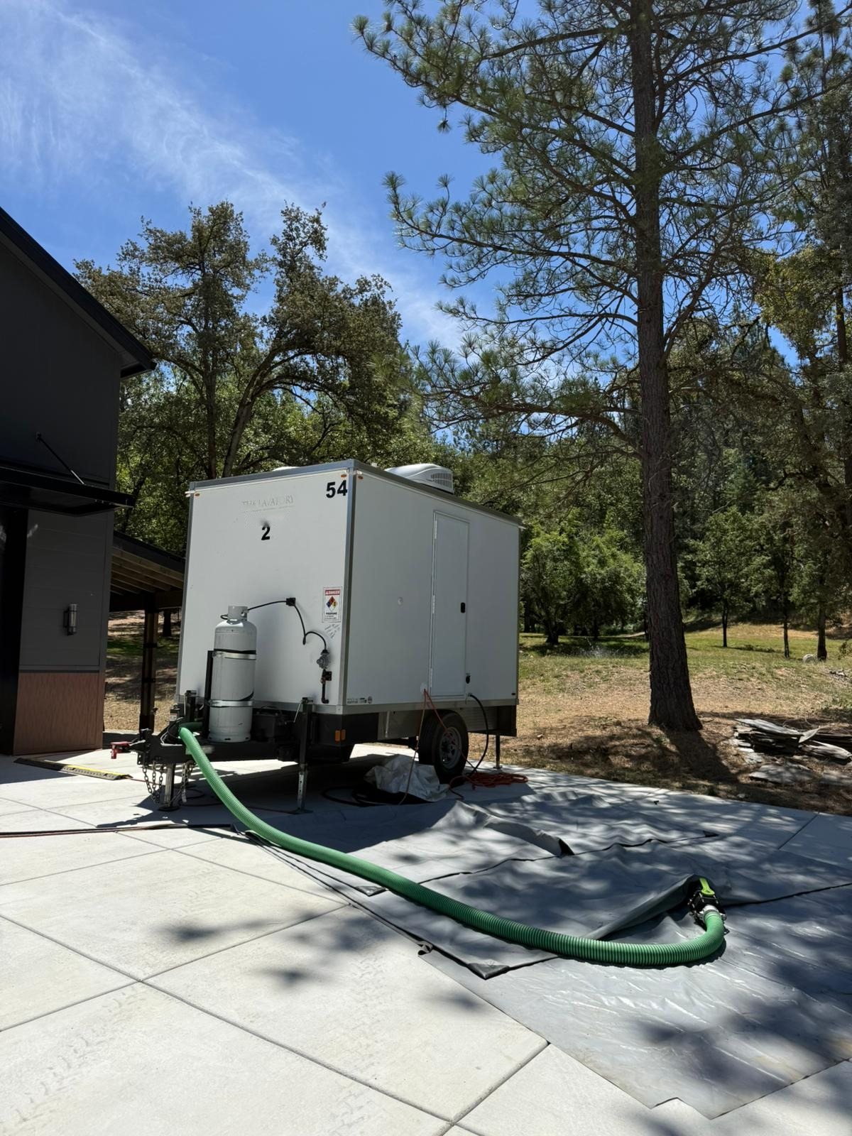 2-station shower and restroom combo trailer with a propane tank and a green sewer hose, set on a new concrete pad under tall pines