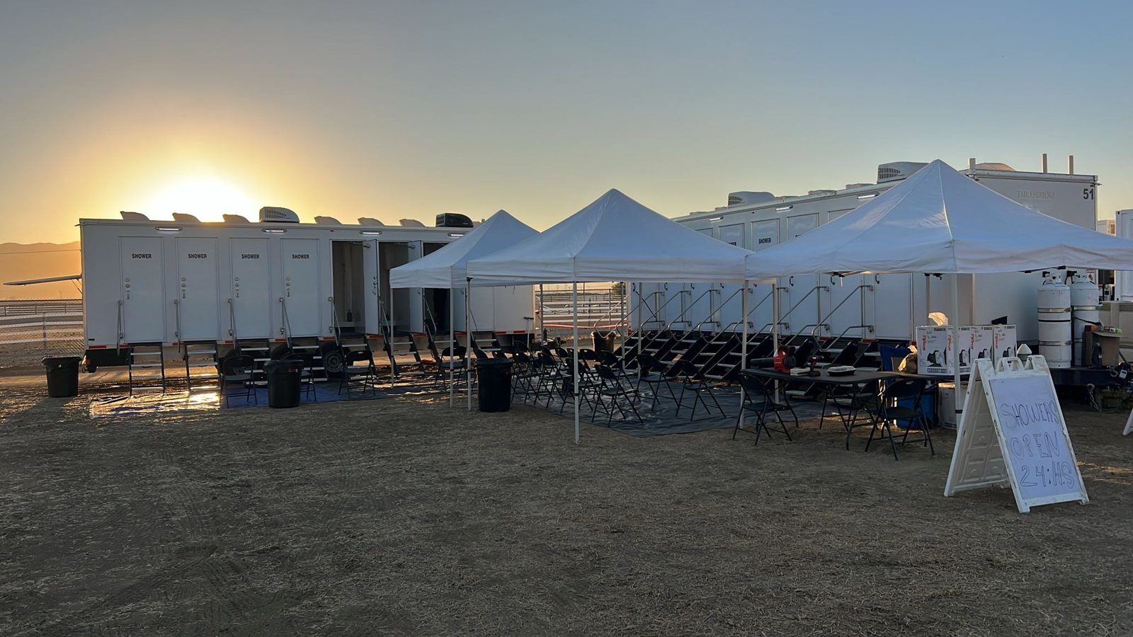 Two 8-station private shower trailers at sunrise on a dirt base camp lot, with white canopies, folding chairs and a sign saying the showers are open 24 hours