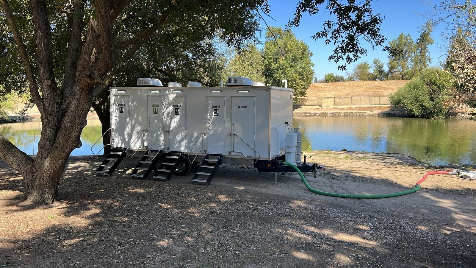 4-station shower, restroom and laundry combo trailer with SHOWER and LAUNDRY doors, parked in oak shade beside a pond with a sewer hose run