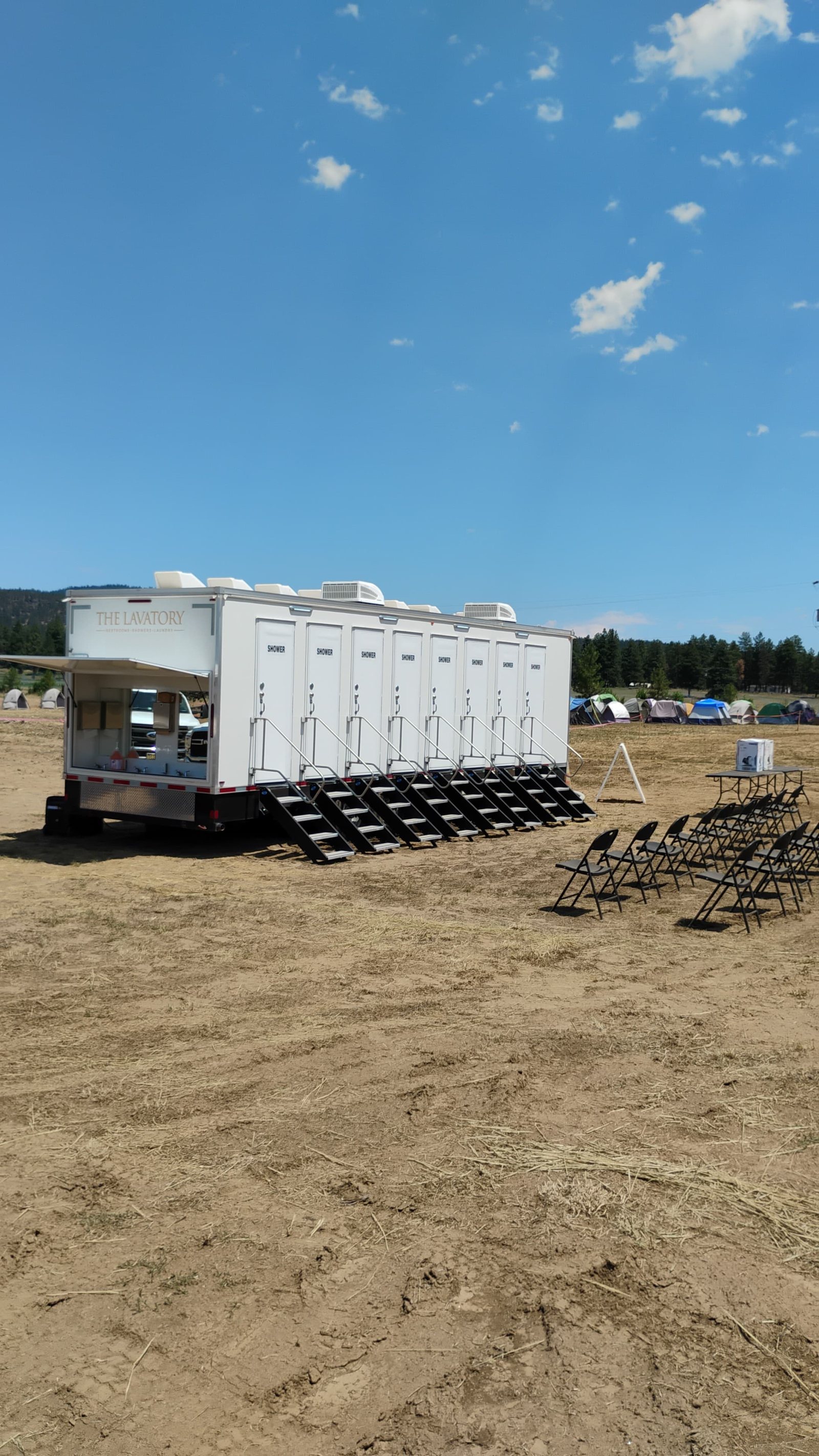 8-station private shower trailer with a stair at each of its eight SHOWER doors and the outside hand wash bank open, set on a dirt field next to a camp of crew tents and folding chairs