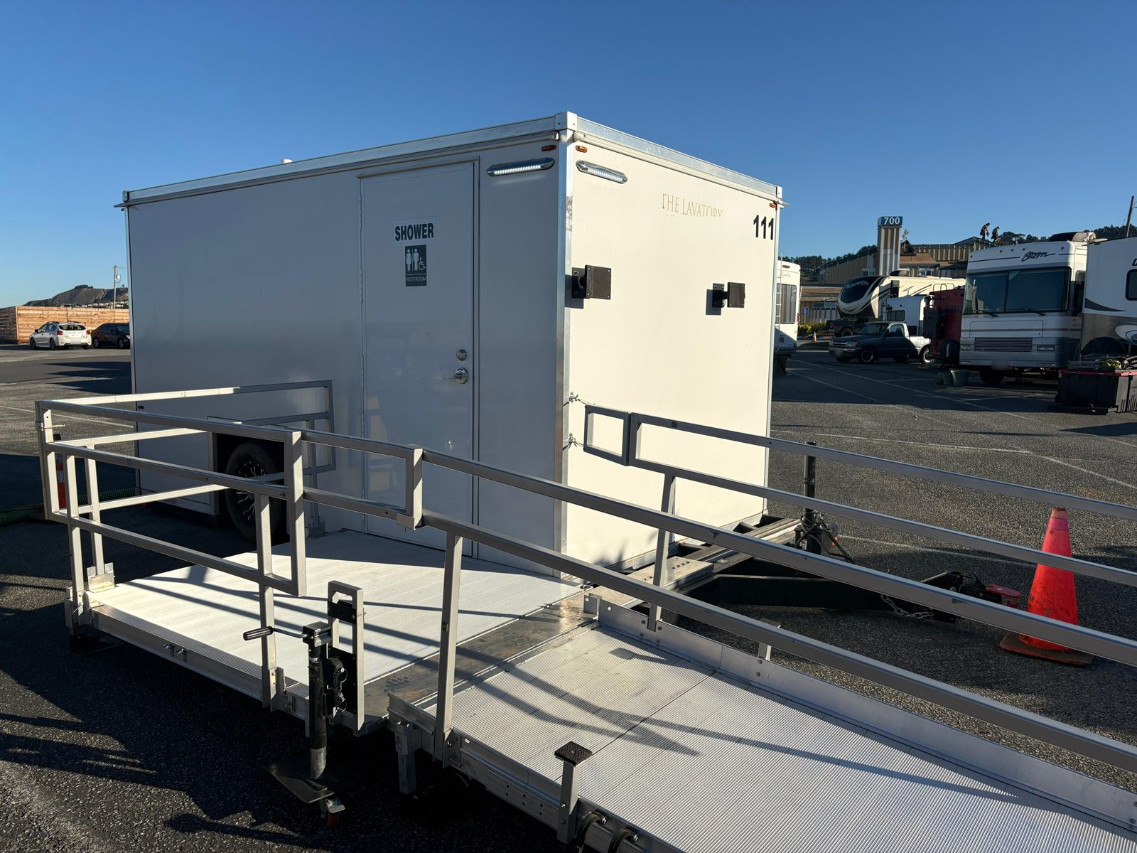 Single-station ADA shower and restroom combo with a long aluminum ramp and rails, parked in a paved lot beside RVs and trailers