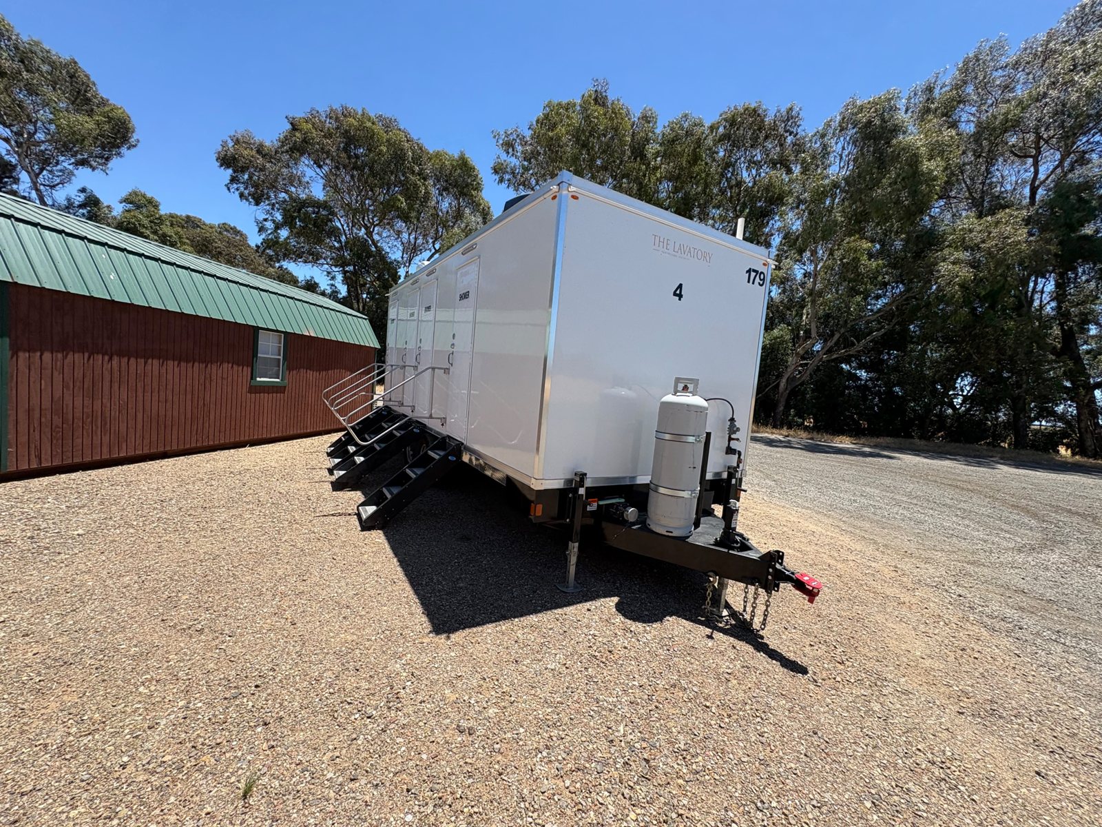 Private shower trailer with four stair units and a propane tank, parked on a gravel lot beside a red wood building with a green metal roof and tall eucalyptus trees