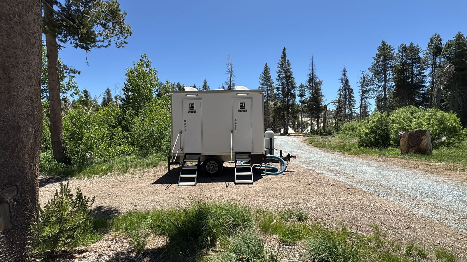 2-station private shower trailer with two stairs, parked off a gravel road among pine trees and green brush